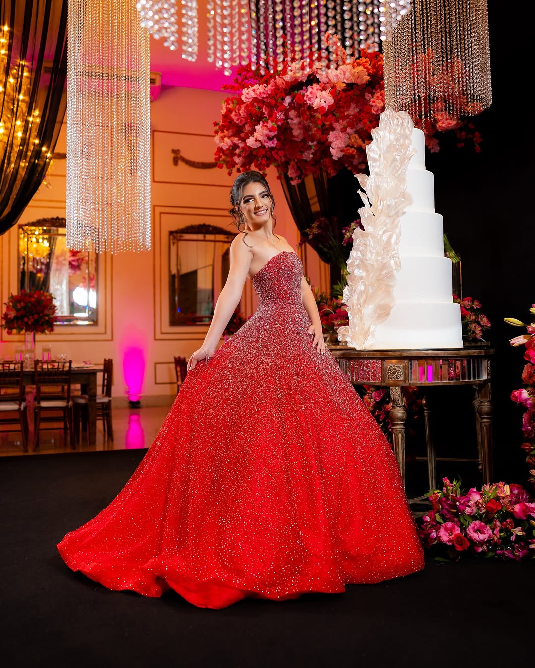 Debutante de vestido vermelho no salão, ao lado de um bolo branco e arranjos de rosas