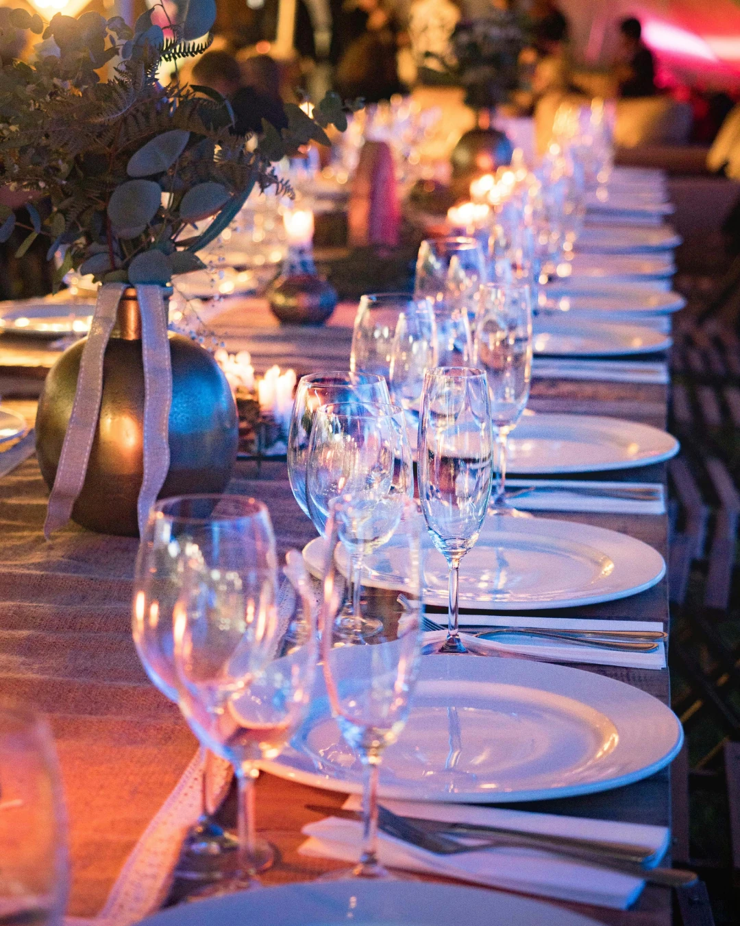 Mesa longa posta com taças de cristal, pratos brancos e velas