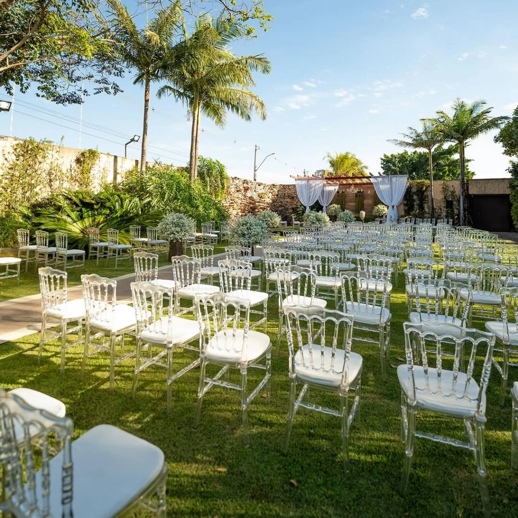 Cadeiras de cristal no gramado do jardim, voltadas para o gazebo com tecidos brancos